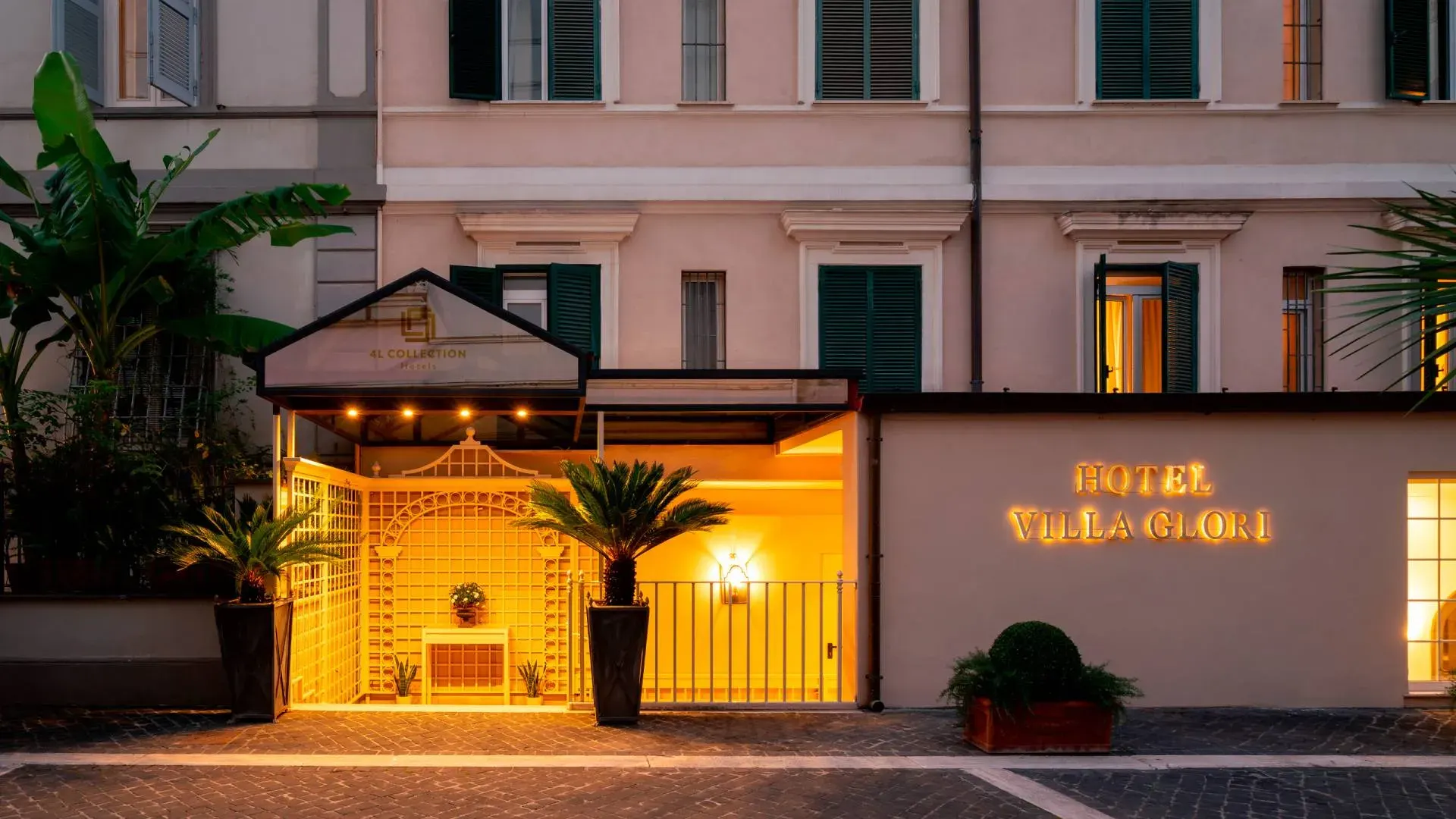 The pink-fronted facade of Hotel Villa Glori at dusk, with a warmly lit entrance framed by palms and the hotel sign beside it.